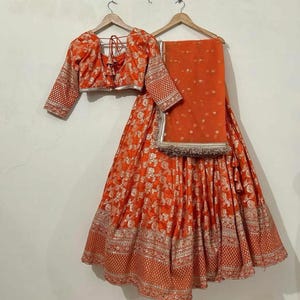 May include: An orange and silver embroidered lehenga with a matching orange blouse. The lehenga has a flared skirt and a fitted bodice. The blouse has a deep neckline and three-quarter sleeves. The outfit is perfect for a special occasion.