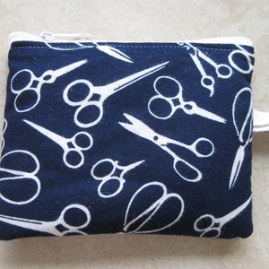 May include: A navy blue zippered pouch with a white cotton strap. The pouch is decorated with a repeating pattern of white scissors.