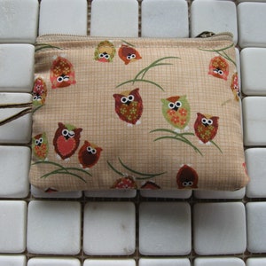 May include: A beige fabric coin purse with a zipper closure. The purse is decorated with a repeating pattern of brown and red owls with white eyes. The owls are surrounded by green grass and brown stems.