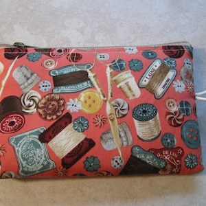 May include: A rectangular, coral-colored fabric pouch with a zipper closure. The pouch is patterned with vintage sewing notions, including spools of thread, buttons, thimbles, and scissors. The design features teal, brown, white, and yellow accents.