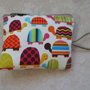 May include: A white zippered pouch with a brown strap. The pouch is decorated with a colorful pattern of turtles with different colored shells and heads.