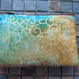 May include: A rectangular, handmade fabric pouch with a swirling, abstract design. The pouch features a color gradient of blue, green, and gold, with copper-colored swirls. It has a brown zipper and trim, and a small loop.