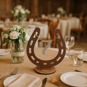 May include: A dark brown wooden horseshoe table number holder featuring a cutout number seven suspended by a thin twine string. The horseshoe is mounted on a circular wooden base with an engraved floral pattern along the bottom edge.