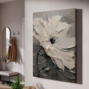Op de afbeelding: Een groot, getextureerd canvas kunstwerk met een witte bloem met een zwart centrum tegen een grijze achtergrond. Het kunstwerk heeft een dikke, impasto-stijl, waardoor een 3D-effect ontstaat. De bloemblaadjes zijn gedetailleerd.