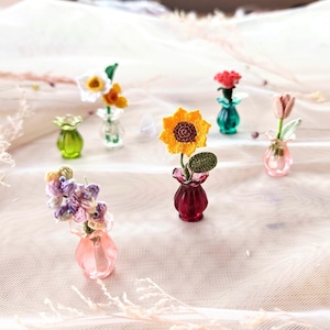 May include: A collection of miniature crochet flowers in small glass vases. The flowers include a yellow sunflower, a red rose, a pink tulip, and other colorful blooms. The vases are in various colors, including green, pink, and red.