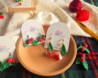 May include: Handmade crochet earrings featuring red berry clusters and green holly leaves. The earrings are displayed on white cards with the "mammi finger" logo, presented on a wooden plate. A crochet necklace is also visible.