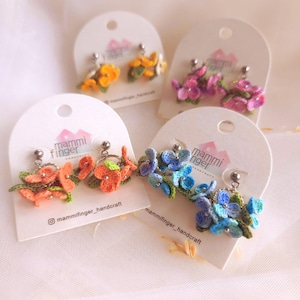 May include: A collection of handmade crochet flower earrings in various colors, including orange, yellow, pink, and blue. Each pair is displayed on a white card with the brand name and social media handle. The earrings feature intricate floral designs.