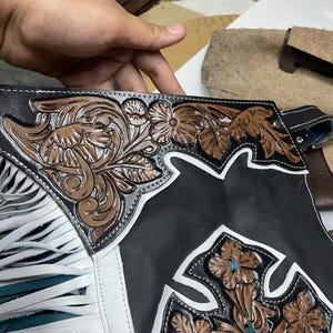 May include: Black leather chaps featuring white leather trim and long white fringe. The surface is decorated with intricate brown floral tooling and a prominent brown leather cross emblem with floral details and a teal leather lace accent.