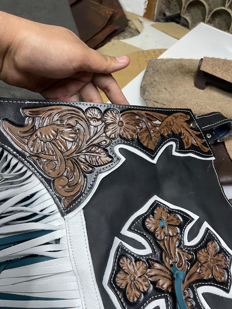 May include: Black leather chaps featuring white leather trim and long white fringe. The surface is decorated with intricate brown floral tooling and a prominent brown leather cross emblem with floral details and a teal leather lace accent.