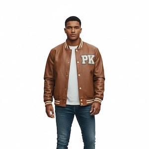 May include: A brown varsity jacket featuring a white and black patch with the letters PK on the chest. The jacket has a snap-button front closure, a ribbed collar, and ribbed cuffs with white stripes, worn over a plain white t-shirt and blue jeans.