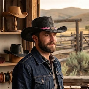 Pode incluir: Um chapéu de cowboy preto com um padrão texturizado semelhante a pele de cobra, apresentando uma faixa trançada rosa brilhante ao redor da copa e um pequeno emblema metálico na frente. O chapéu tem uma aba larga e curva e copa estruturada.
