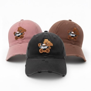 May include: Three baseball caps in pink, black, and brown. Each cap features a distressed design and an embroidered teddy bear holding a sign that reads "BROKEN".