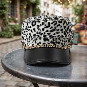 May include: A military-style cap featuring a white and black leopard print faux fur crown. The cap includes a smooth black brim and a decorative gold-tone metal chain detail that runs across the front, secured by two gold-tone circular side buttons.