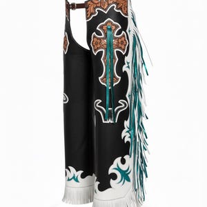 May include: Black leather chaps featuring a brown floral tooled leather waistband and upper yoke. The legs are decorated with a large brown tooled leather cross, teal ribbon accents, white leather overlays, and long white and teal fringe trim.