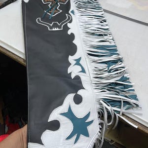 May include: A black leather chap featuring a white leather overlay with teal accents. The design includes a decorative cross emblem at the top, a bull horn cutout, and long white fringe along the side. The piece is constructed with white stitching.
