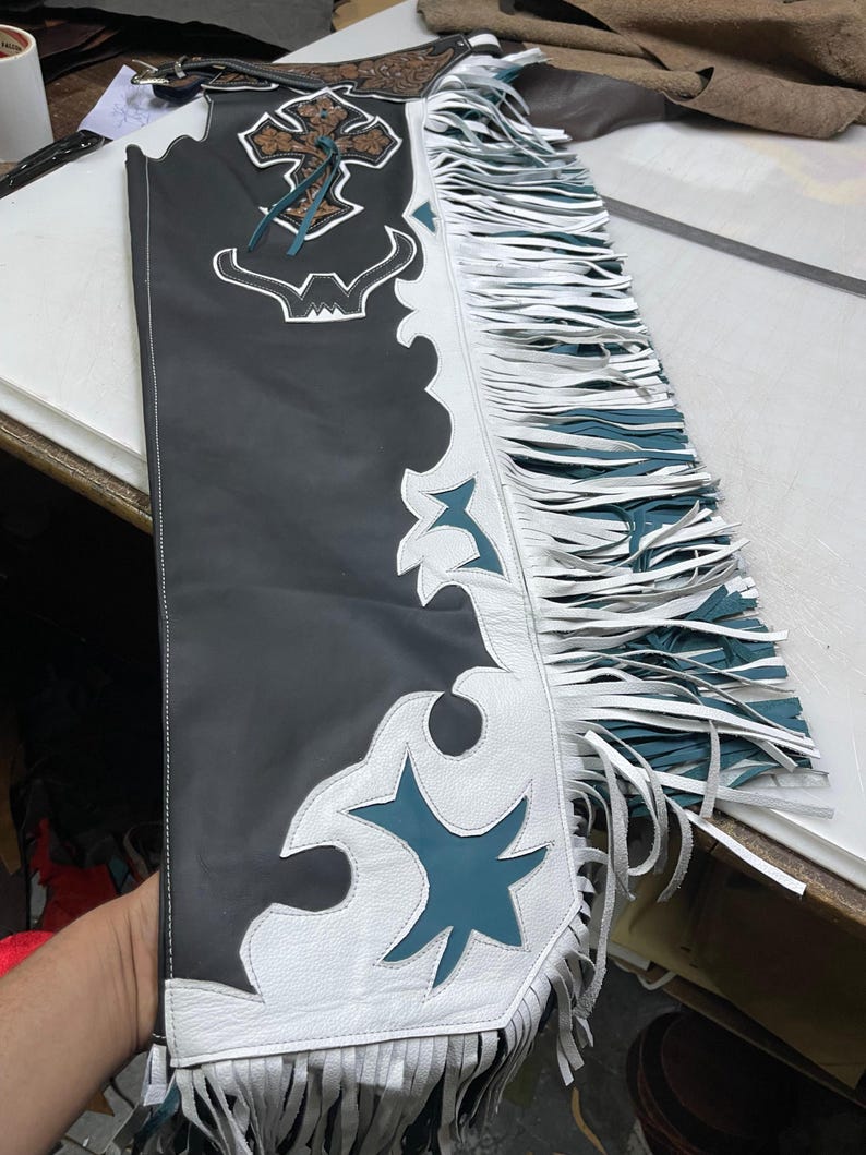 May include: A black leather chap featuring a white leather overlay with teal accents. The design includes a decorative cross emblem at the top, a bull horn cutout, and long white fringe along the side. The piece is constructed with white stitching.