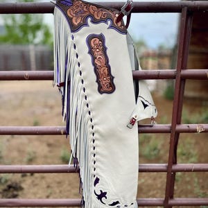 Op de afbeelding: Off-white leren chaps met gedetailleerd bruin bloemmotief en paarse accenten. Het kledingstuk is voorzien van lange witte franjes aan de zijkant, paarse leren biezen en zilverkleurige metalen gespen voor verstelling en bevestiging.