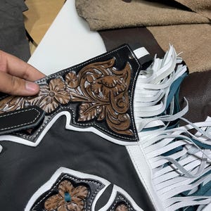 May include: Black leather chaps featuring brown floral embossed leather panels, white leather trim, and long white fringe accents. The design includes a cross-shaped cutout detail with intricate floral tooling and white stitching throughout the construction.
