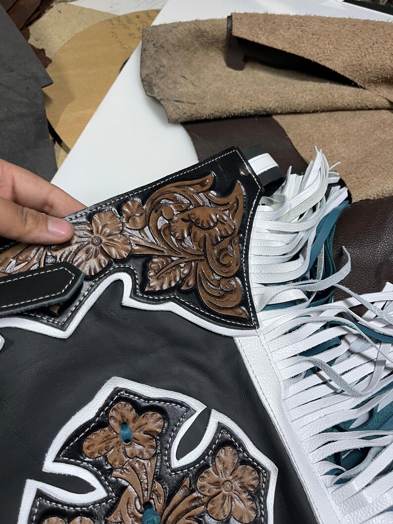 May include: Black leather chaps featuring brown floral embossed leather panels, white leather trim, and long white fringe accents. The design includes a cross-shaped cutout detail with intricate floral tooling and white stitching throughout the construction.