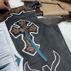 May include: Black leather chaps featuring white fringe trim, a large embossed brown leather cross with floral detailing, and a turquoise leather lace accent. The design includes white leather piping and a stylized white bull horn graphic at the base.