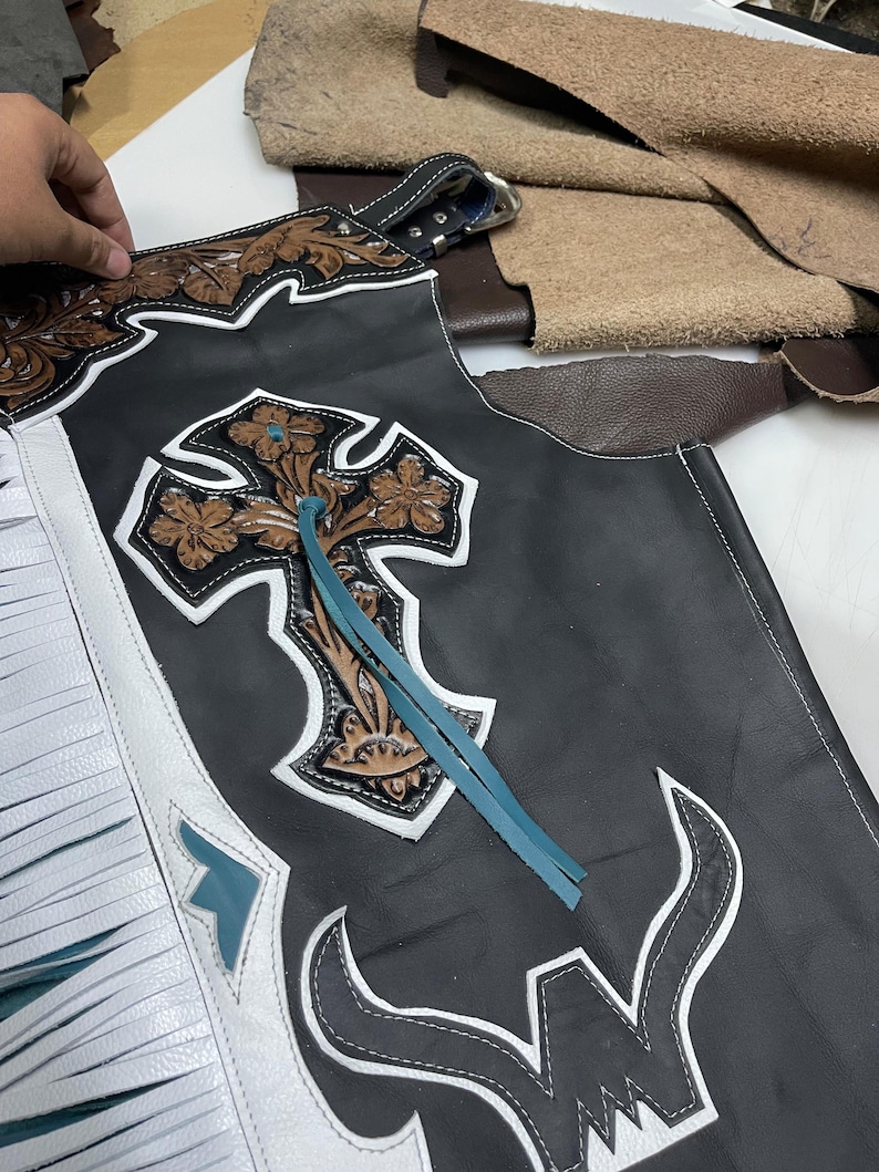 May include: Black leather chaps featuring white fringe trim, a large embossed brown leather cross with floral detailing, and a turquoise leather lace accent. The design includes white leather piping and a stylized white bull horn graphic at the base.