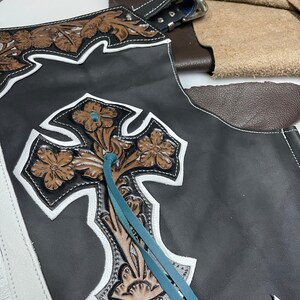 May include: Black leather chaps featuring a large, embossed brown floral cross design with white leather trim. The piece includes turquoise leather lacing and a silver-toned metal buckle at the top, showcasing intricate tooling and stitching details.