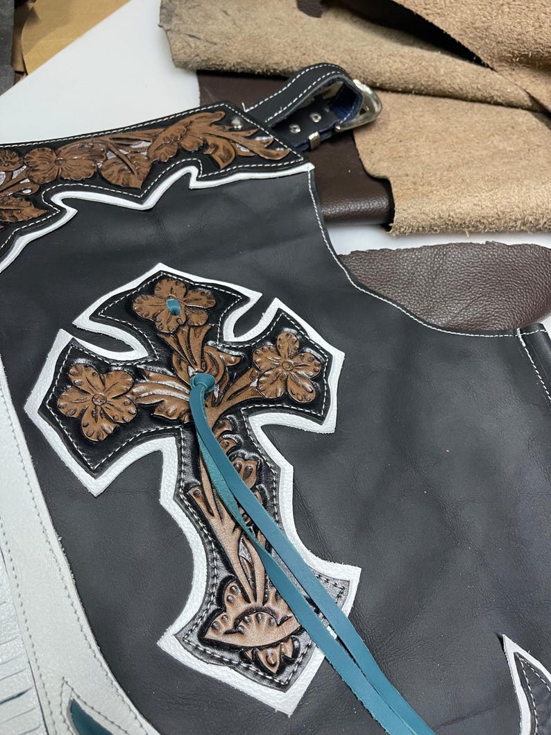 May include: Black leather chaps featuring a large, embossed brown floral cross design with white leather trim. The piece includes turquoise leather lacing and a silver-toned metal buckle at the top, showcasing intricate tooling and stitching details.