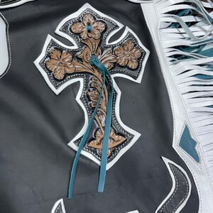 May include: Black leather chaps featuring a large, embossed brown floral cross appliqu&eacute; with white leather trim. The design includes long white and teal leather fringe along the side, intricate floral tooling, and teal leather lacing accents.