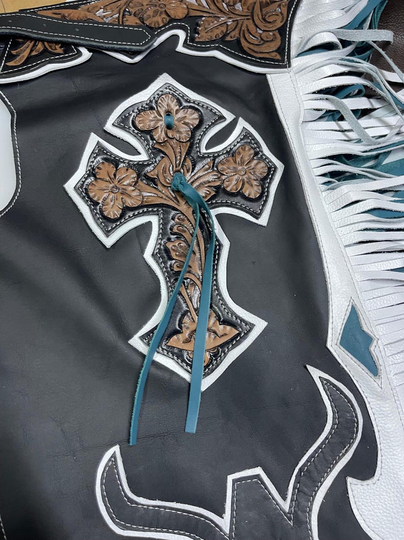 May include: Black leather chaps featuring a large, embossed brown floral cross appliqu&eacute; with white leather trim. The design includes long white and teal leather fringe along the side, intricate floral tooling, and teal leather lacing accents.