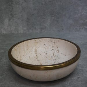 May include: A round, off-white bowl with a brass-colored rim. The interior of the bowl has a textured appearance with small dark spots. The bowl is set against a gray background.