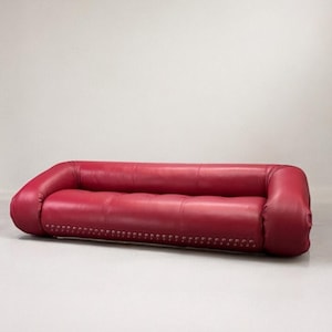 Könnte beinhalten: Ein rotes Ledersofa mit niedriger Form und abgerundeten Armlehnen. Das Sofa hat getuftete Kissen und eine Reihe von Ziernieten an der Basis. Das Sofa ist etwa 200 cm lang. Ein modernes Möbelstück.