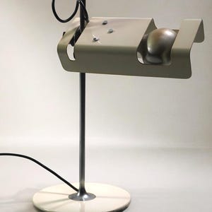 Può includere: Una lampada da scrivania moderna e minimalista con un paralume color crema e una base in metallo color argento. La lampada presenta una sorgente luminosa sferica e un dettaglio di filo nero curvo. La base è di forma circolare e la lampada è collegata a un cavo di alimentazione nero.