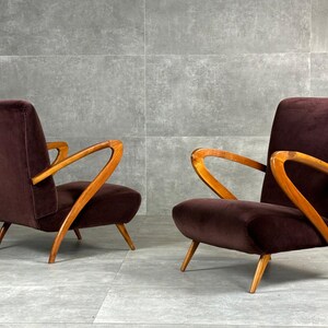 May include: Two mid-century modern armchairs. The chairs have a dark burgundy velvet upholstery and curved wooden armrests and legs. The chairs are set against a gray background.