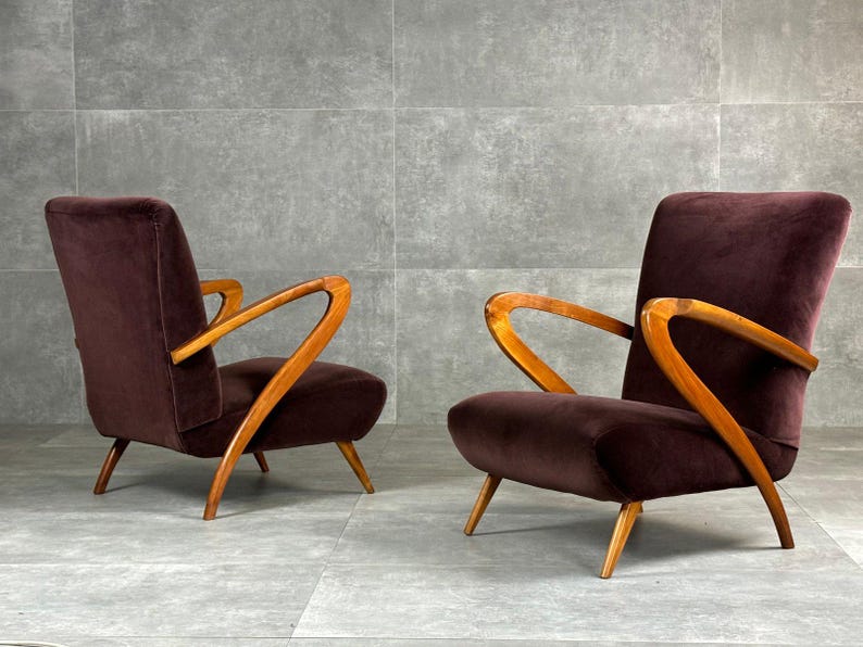 May include: Two mid-century modern armchairs. The chairs have a dark burgundy velvet upholstery and curved wooden armrests and legs. The chairs are set against a gray background.