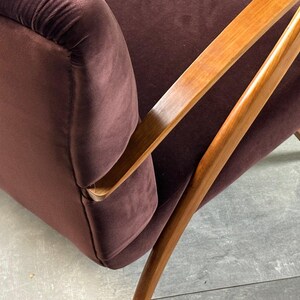 May include: Close-up of a mid-century modern armchair. The chair features a deep burgundy velvet upholstery and a wooden frame with angled armrests and legs. The wood has a warm, natural finish.