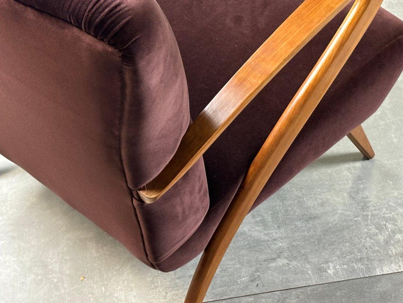 May include: Close-up of a mid-century modern armchair. The chair features a deep burgundy velvet upholstery and a wooden frame with angled armrests and legs. The wood has a warm, natural finish.