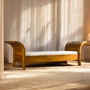 Può includere: Una panca in legno con un cuscino colorato chiaro. La panca ha braccioli curvi e una calda finitura in legno naturale. La panca è posizionata su un tappeto beige in una stanza con una tenda bianca.