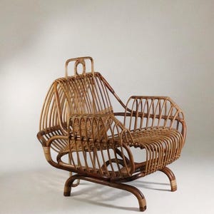 Puede incluir: Una silla vintage de ratán con un diseño único. La silla presenta un respaldo curvo, reposabrazos y un asiento de ratán tejido. El marco es de madera marrón oscuro. La silla tiene un asa en la parte superior del respaldo.