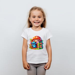 May include: A white t-shirt with a blue cartoon character standing in front of a red and white mushroom house. The character has blue ears and is holding a red object. There are smaller mushrooms in the background.