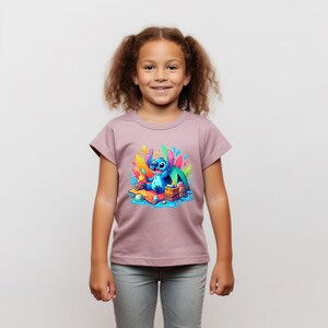 Puede incluir: Una camiseta de color malva con un gr&aacute;fico colorido de un personaje de dibujos animados azul. El personaje est&aacute; sentado sobre una superficie de madera con un fondo colorido. La camiseta la lleva una ni&ntilde;a con el pelo casta&ntilde;o rizado y pantalones grises.