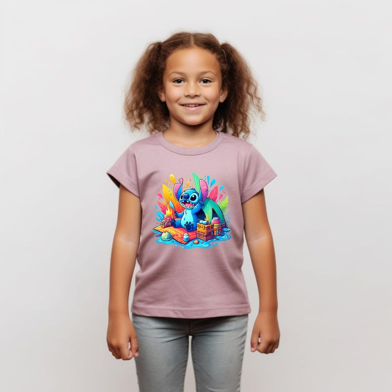 Puede incluir: Una camiseta de color malva con un gr&aacute;fico colorido de un personaje de dibujos animados azul. El personaje est&aacute; sentado sobre una superficie de madera con un fondo colorido. La camiseta la lleva una ni&ntilde;a con el pelo casta&ntilde;o rizado y pantalones grises.