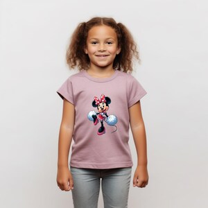 May include: A young girl wearing a mauve t-shirt with a graphic of Minnie Mouse holding pom-poms. The graphic is in a cartoon style and features Minnie Mouse in a red and white polka dot dress with a red bow.
