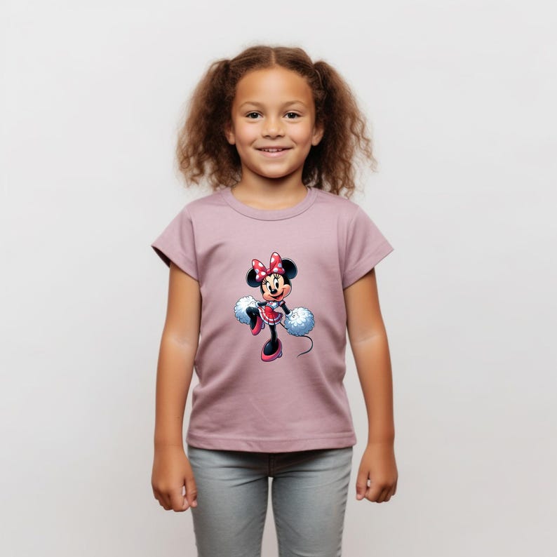 May include: A young girl wearing a mauve t-shirt with a graphic of Minnie Mouse holding pom-poms. The graphic is in a cartoon style and features Minnie Mouse in a red and white polka dot dress with a red bow.