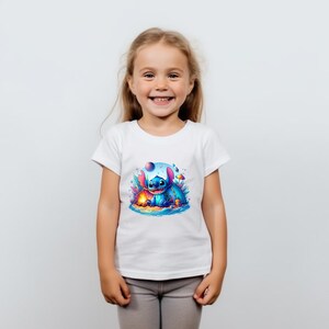Puede incluir: Una camiseta blanca con un gr&aacute;fico colorido de un personaje de dibujos animados azul con orejas grandes, sentado en una playa con una fogata. El personaje est&aacute; rodeado de detalles vibrantes y caprichosos. La camiseta la lleva una ni&ntilde;a.