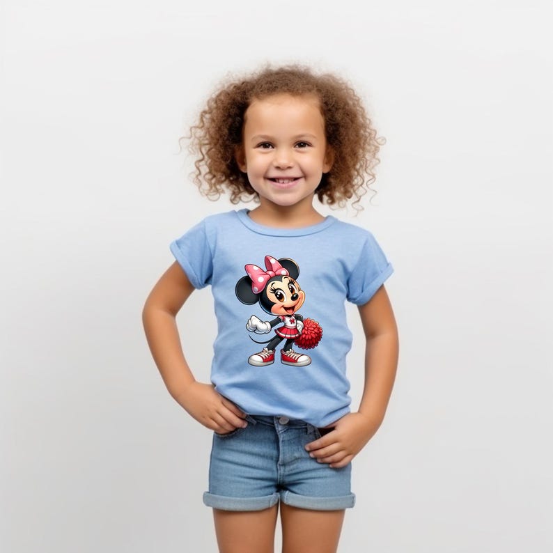 May include: A young girl wearing a light blue t-shirt with a cartoon image of Minnie Mouse dressed as a cheerleader. Minnie Mouse is holding a red pom-pom and has a red skirt and white shoes.
