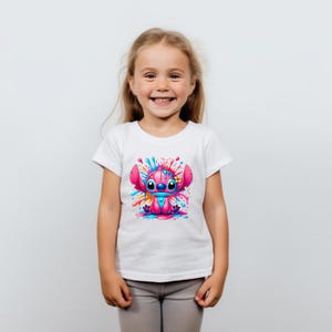May include: A white t-shirt featuring a pink cartoon character with blue eyes and a flower on its head. The character is surrounded by colourful paint splatters.