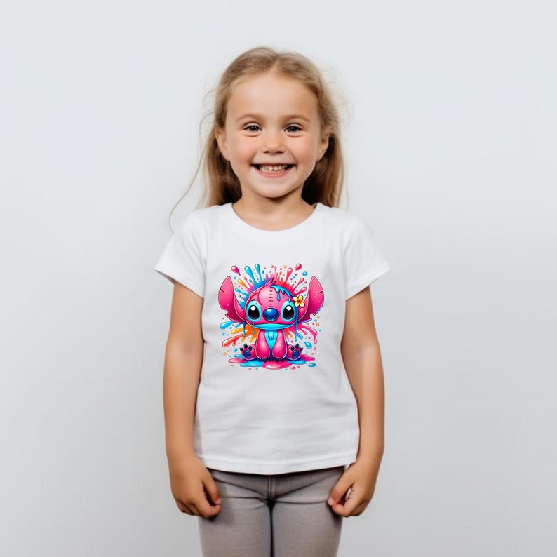 May include: A white t-shirt featuring a pink cartoon character with blue eyes and a flower on its head. The character is surrounded by colourful paint splatters.