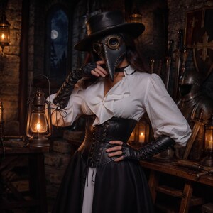 May include: A plague doctor mask with brass-rimmed eye lenses, a black wide-brimmed hat, a white ruffled blouse, a black lace-up corset, and long black leather gloves. A metal lantern with a glowing light is held in one hand, illuminating the scene.