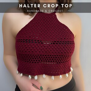 May include: A burgundy halter crop top, handmade and crocheted. The top features a detailed openwork design with a row of horizontal openings. The bottom edge is adorned with small, dangling shell embellishments. The text "HALTER CROP TOP HANDMADE & CROCHET" is at the top.