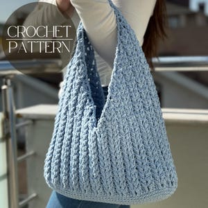 May include: A light blue crochet bag with a textured stitch pattern. The bag has a wide opening and a long strap. The words "CROCHET PATTERN" are in white text on the upper left side of the image.